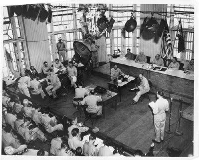 A black and white photo, wide shot from above of approximately 20 men wearing jumpsuits, mostly sitting, and some standing. A makeshift courtroom with wooden tables which five men are sitting at, and court reporters appearing to type on a typewriter.