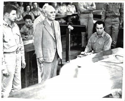A black and white photo of three men in the foreground, two Caucasian and the middle appearing to be Asian. The caucasian men wear military suits and the Asian man wears a suit and tie. People in the background are blurred, but sit as if in a courtroom.