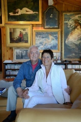 A white male and female couple in their 70s sitting on a couch surrounded by fine art paintings in their home.