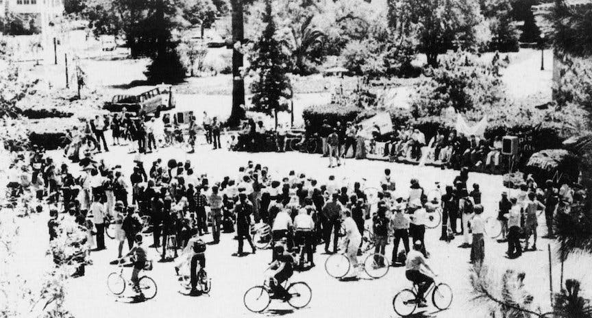 A large crowd, including individuals on bicycles, is gathered to watch the protest.