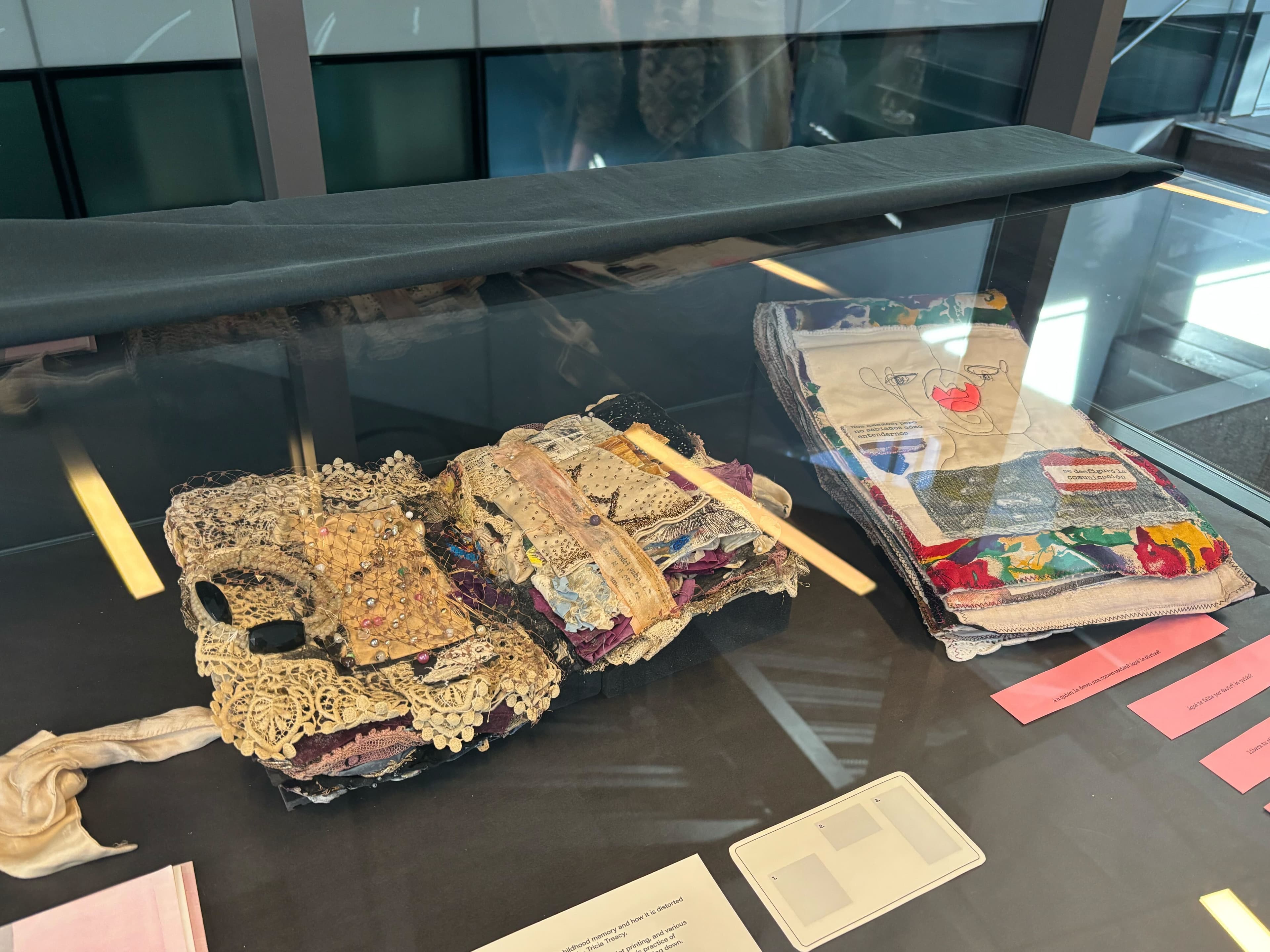 Two pieces of multimedia art in a glass display. The piece on the left has layered fabric pieces with lace and various textiles. The piece on the right is an open book made of sewn fabrics.