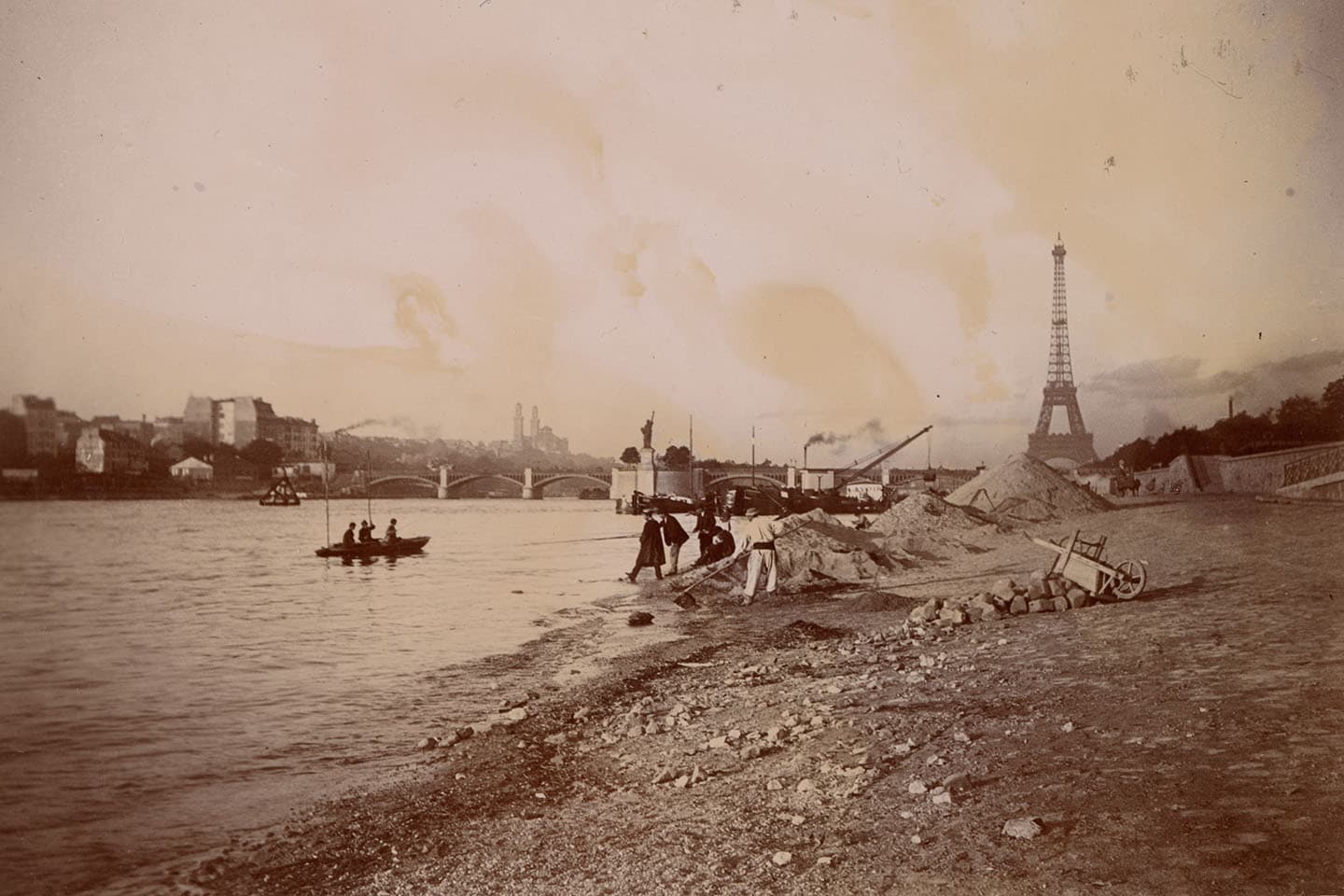 Group of people on the riverbank of the Seine River in Paris with the Eiffel Tower and a replica of the Statue of Liberty in the background. 