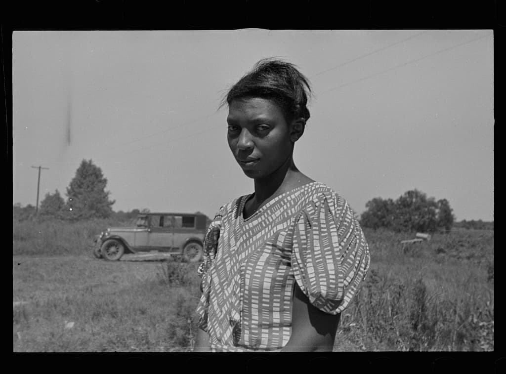  Half-length photo of a Black woman looking into the camera, standing in a field with a 1930’s-era car in the background.