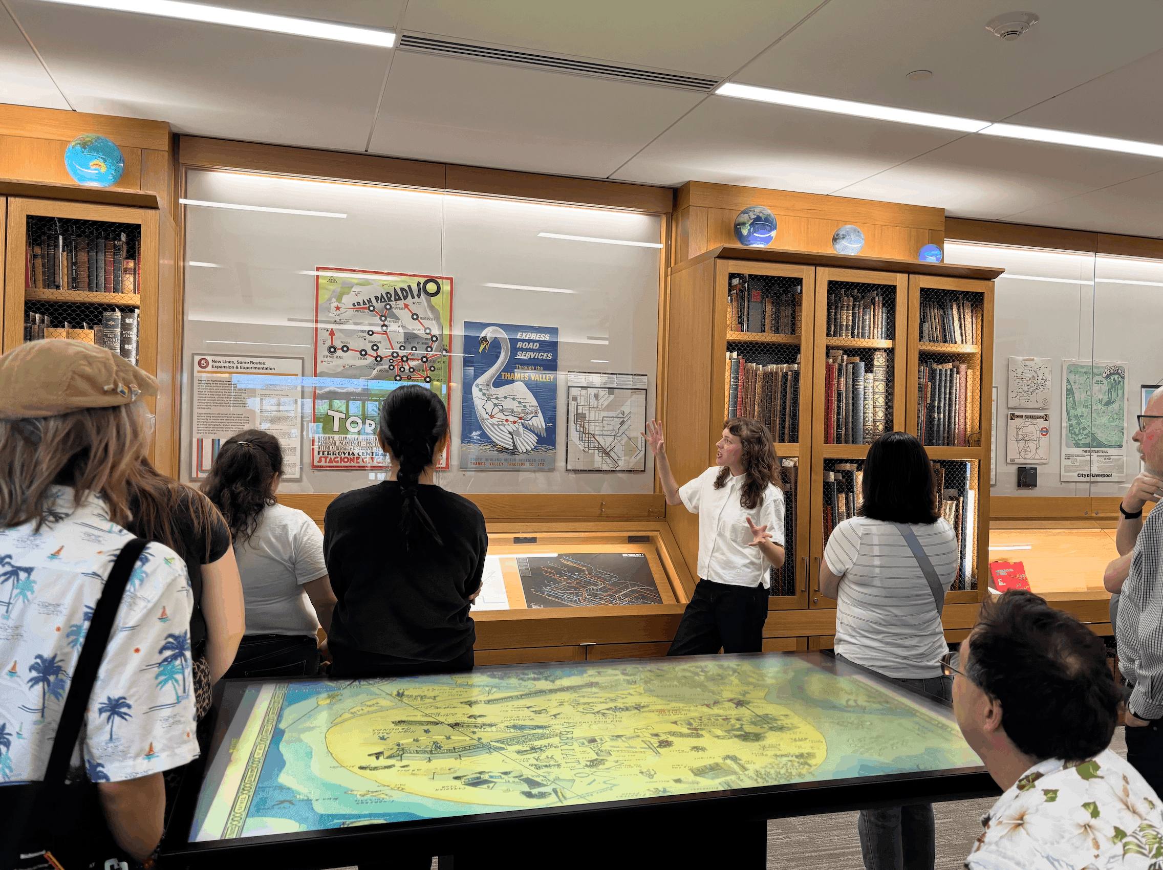 A group of exhibit goers are gathered around a display, listening to a presenter talk about a map on display.