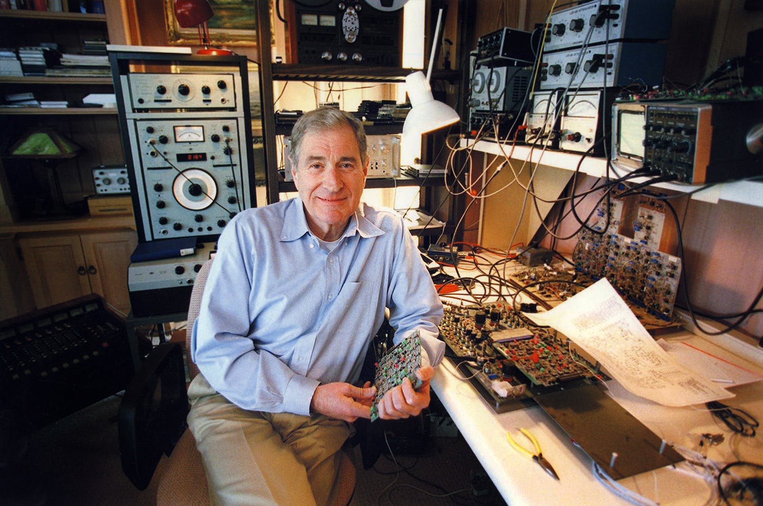 Ray Dolby seated and smiling holding a device at his home workbench surrounded by audio equipment