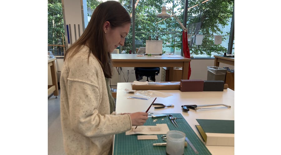 Abigail Mead standing at a desk  working on a copy of a 19th century stationary binding using conservation tools.