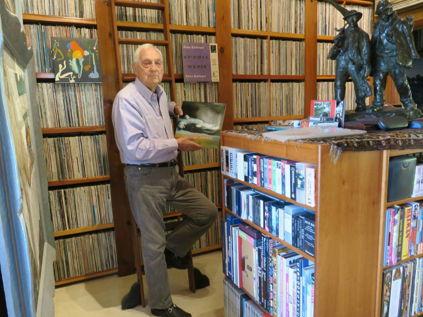 White male standing in front of his album collection with his knee bent on ladder holding an album. 