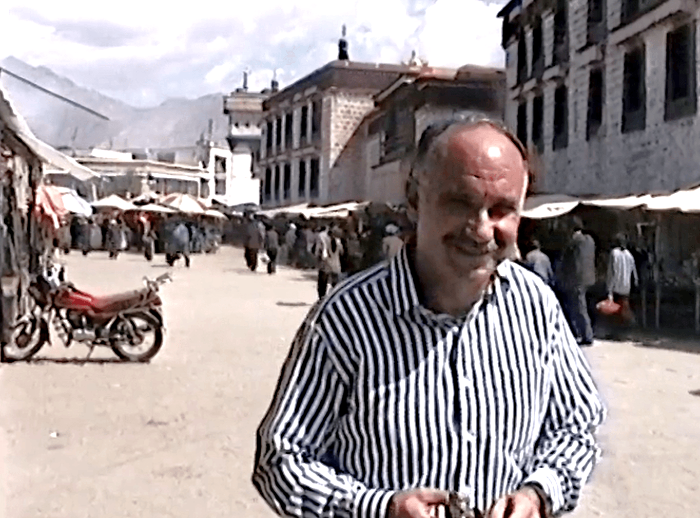 A man in a striped shirt smiles on a bustling market street with distant mountains under a partly cloudy sky.