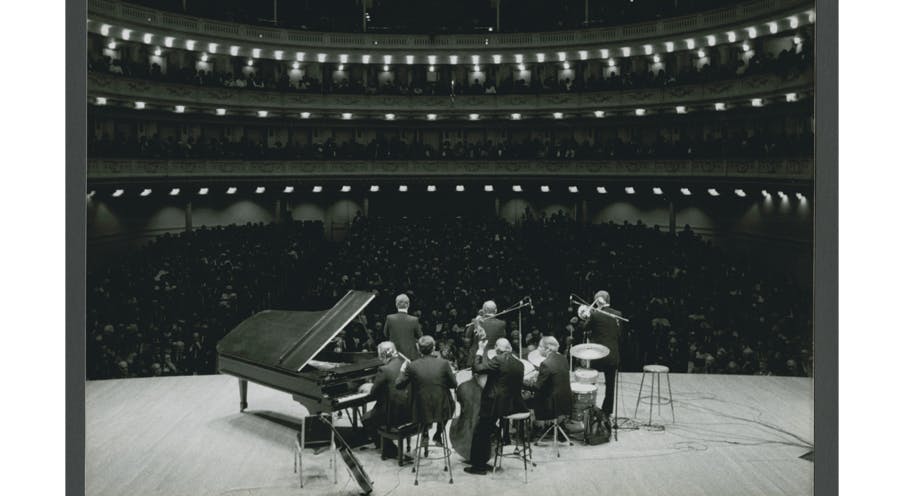 A black and white photo of a jazz band with musical instruments on a stage facing a large audience.