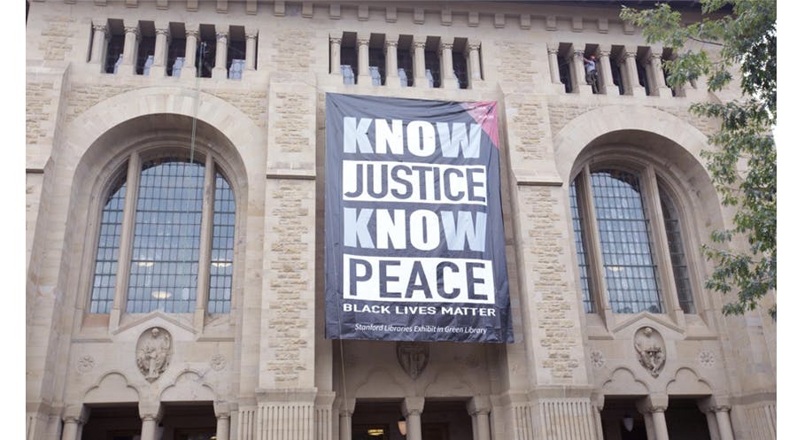 A large black and white banner, with words Know Justice Know Peace, Black Lives Matter, hung on the facade of Green Library.