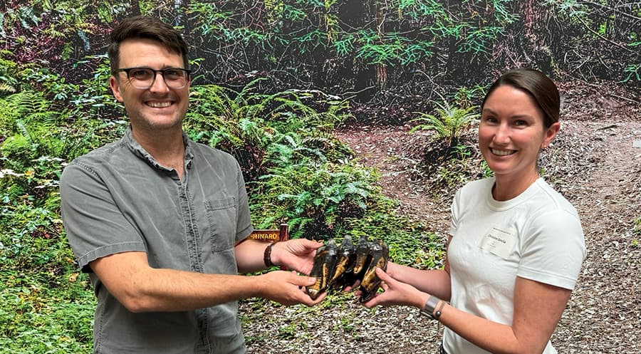 Two people standing in front of a woodsy backdrop holding a giant tooth from a Mastodon.