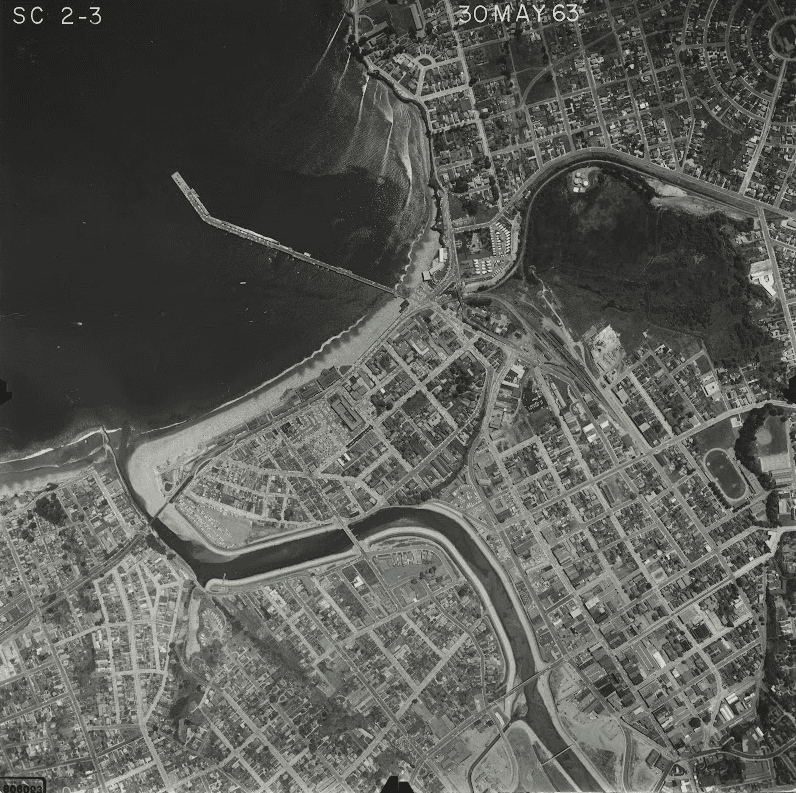 Black-and-white aerial view of a coastal city with a pier and river winding through.