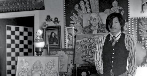 Martin Wong surrounded by framed artwork standing and holding a hat