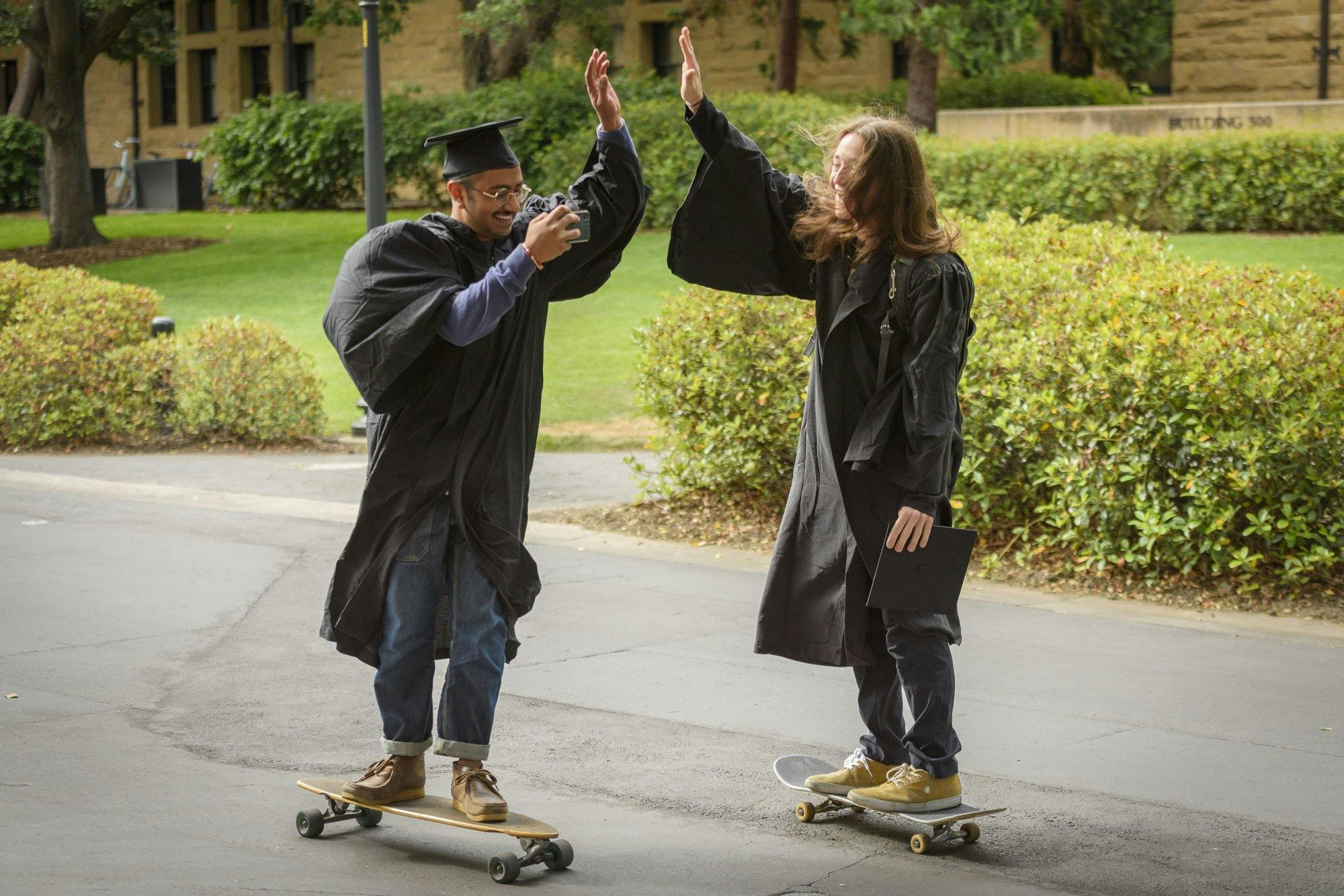 Two Stanford graduates skate board in cap and gown