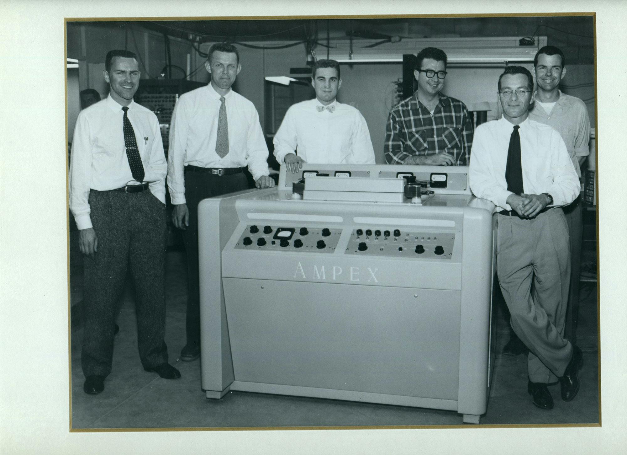Six men standing behind the first AMPEX Video Tape Recorder device. The third man from the left wearing a bow tie is Ray Dolby. The photo is black and white.