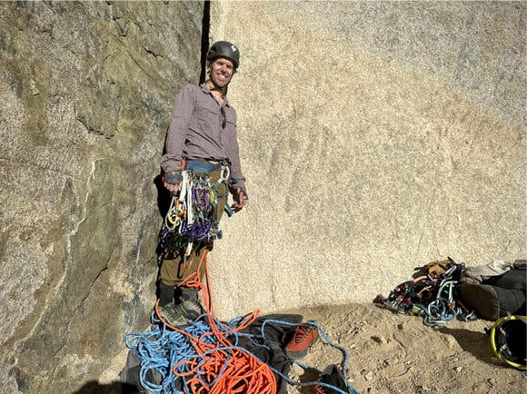 A man stands smiling in front of a boulder with rock climbing gear around his waste and several ropes on the ground.