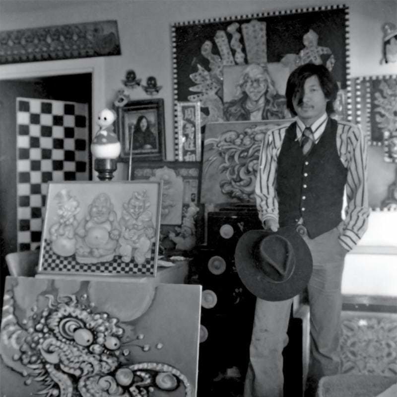 Martin Wong surrounded by framed artwork, standing with a hat in his hand